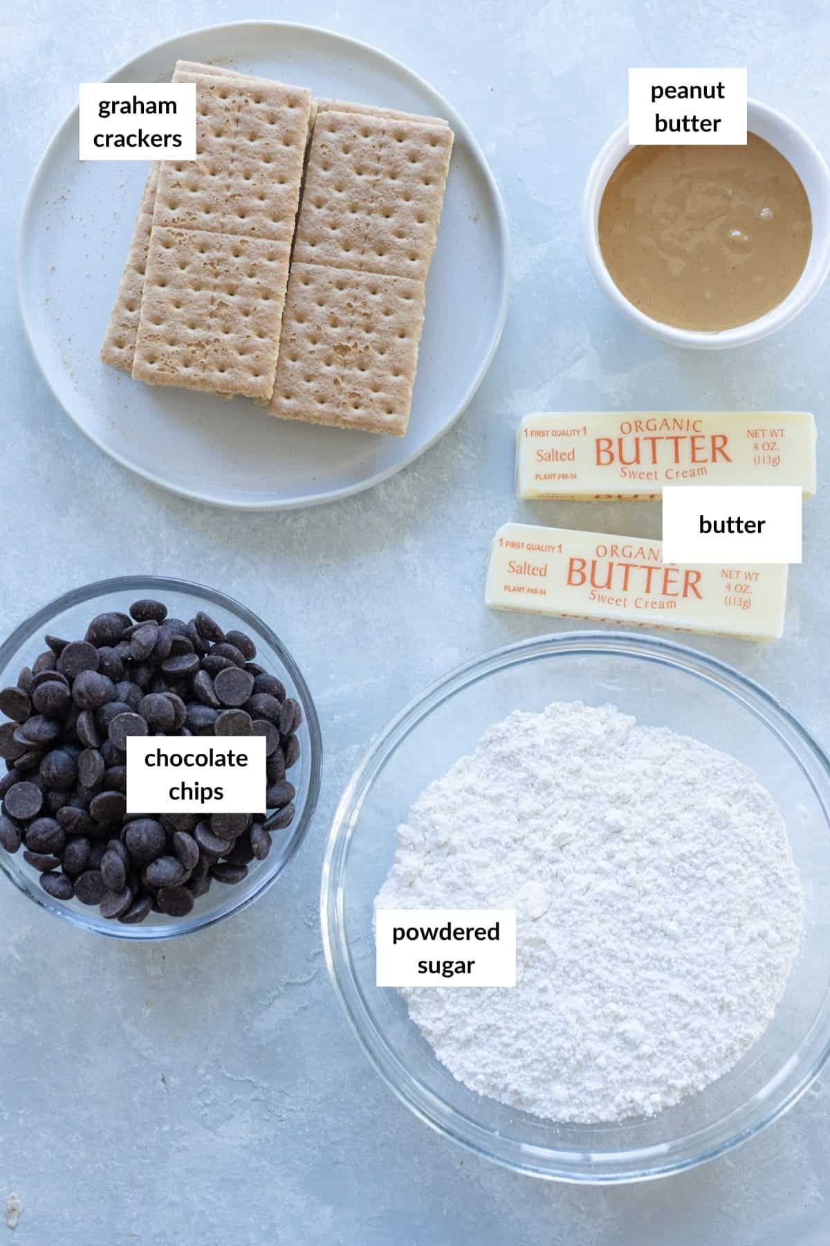 Five labeled ingredients on a countertop for No Bake Buckeye Bars: graham crackers, peanut butter, butter, chocolate chips, and powdered sugar, each in a separate bowl or plate.