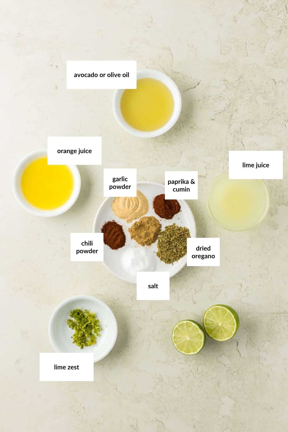A variety of labeled ingredients in bowls and on a plate, including oils, juices, spices, salt, and lime zest&mdash;perfect for making a flavorful chicken fajita marinade&mdash;are arranged on a light surface.