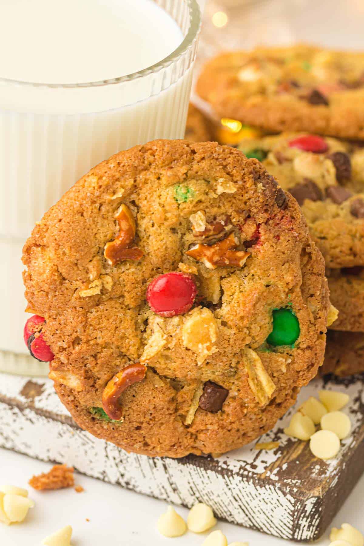 A close-up of a loaded kitchen sink cookie with colorful candy-coated chocolates, pretzel pieces, and chocolate chips, resting against a glass of milk.