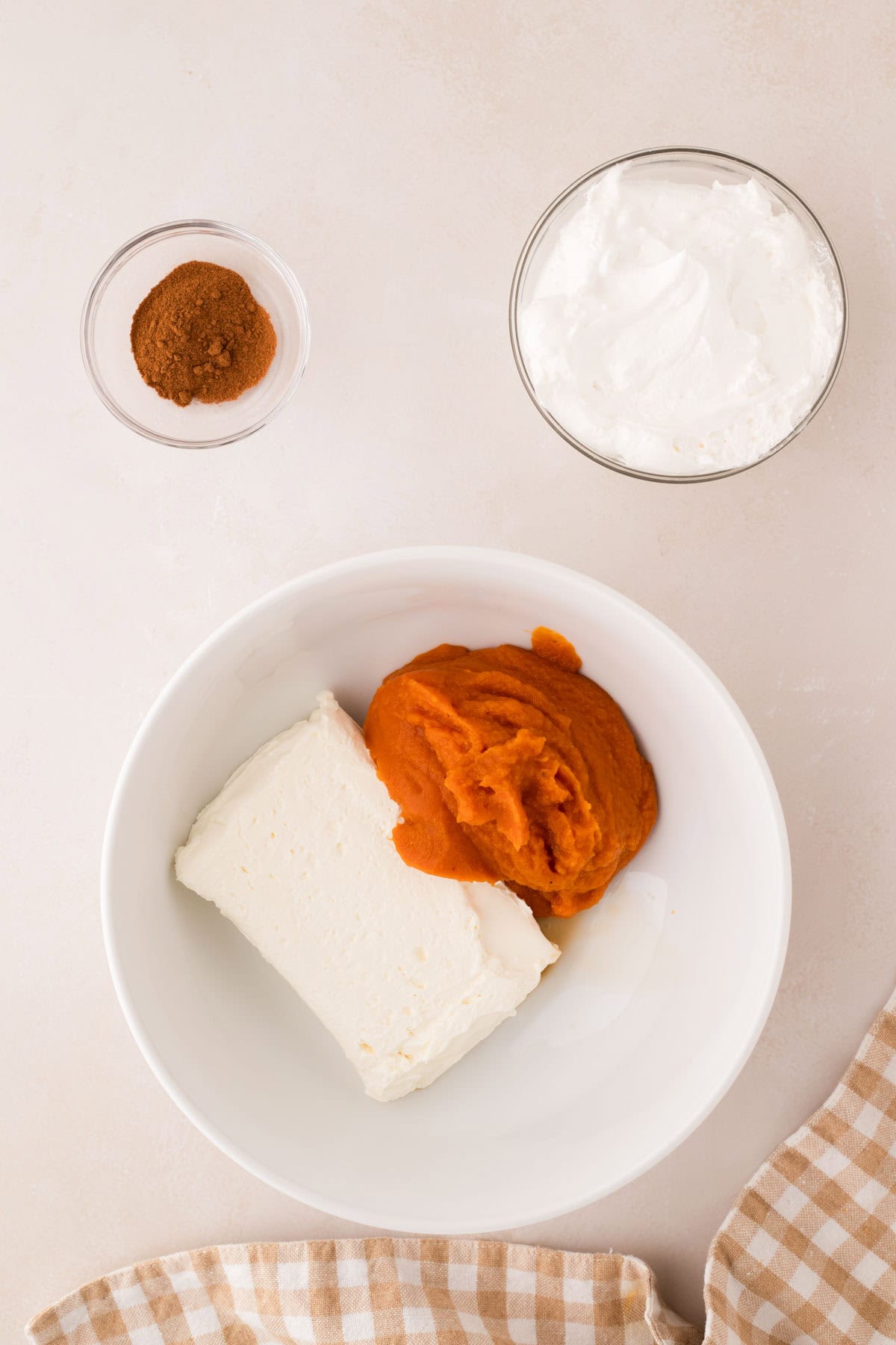 A white bowl with cream cheese and pumpkin puree, next to small bowls of whipped topping and spices, on a light surface with a checkered cloth.
