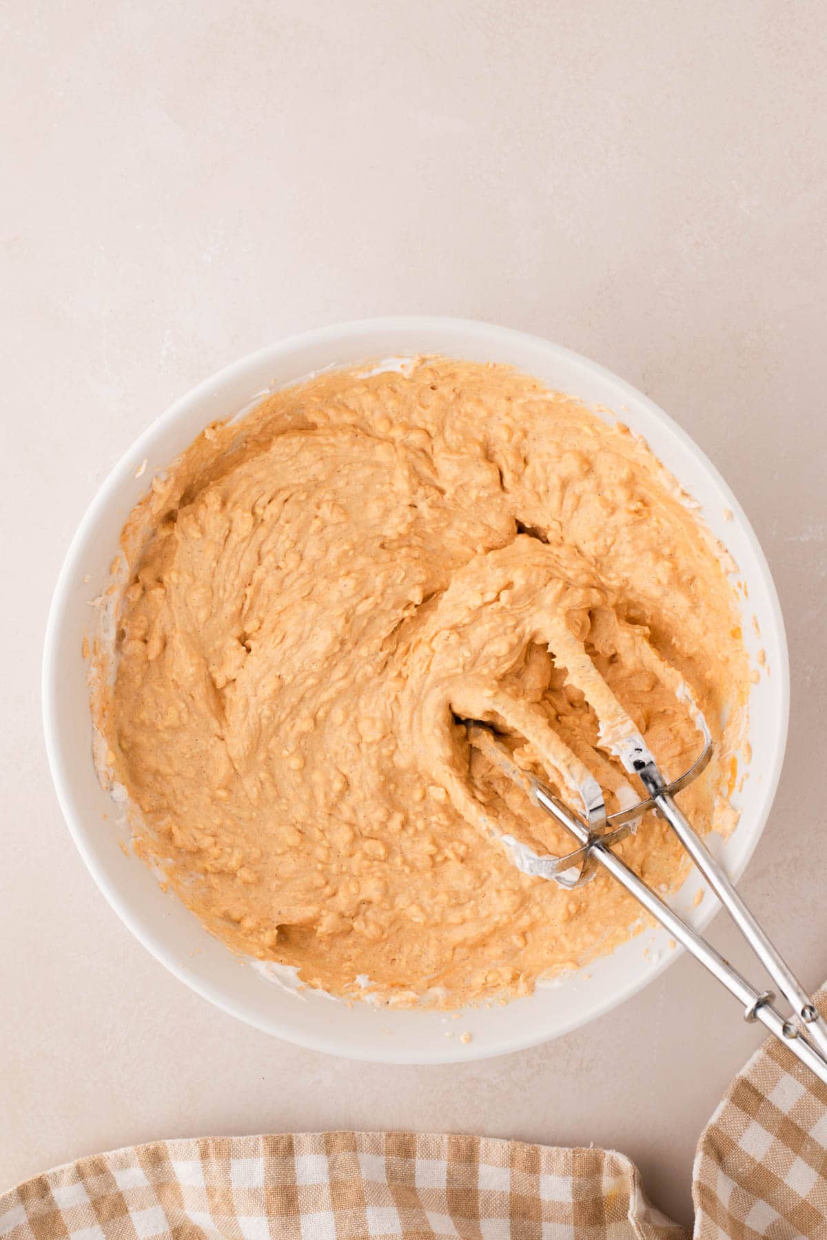 A white bowl filled with creamy dip mix and two metal beaters resting inside, placed on a beige surface next to a checkered cloth.