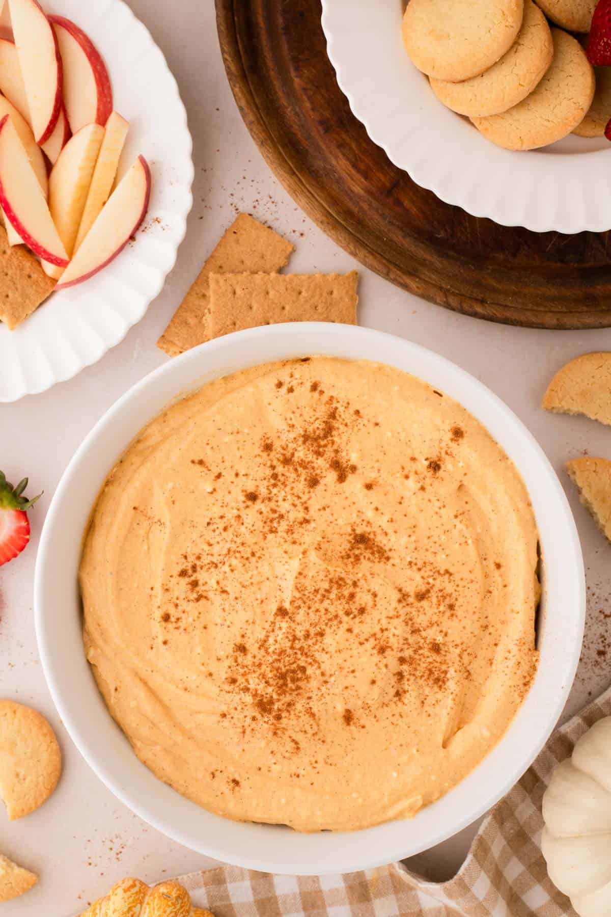 A bowl of creamy pumpkin pie dip sprinkled with cinnamon, surrounded by sliced apples, cookies, graham crackers, and strawberries on plates.