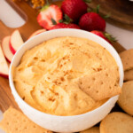 A bowl of pumpkin pie dip topped with a sprinkle of cinnamon, surrounded by graham crackers, cookies, apple slices, and strawberries on a wooden board.