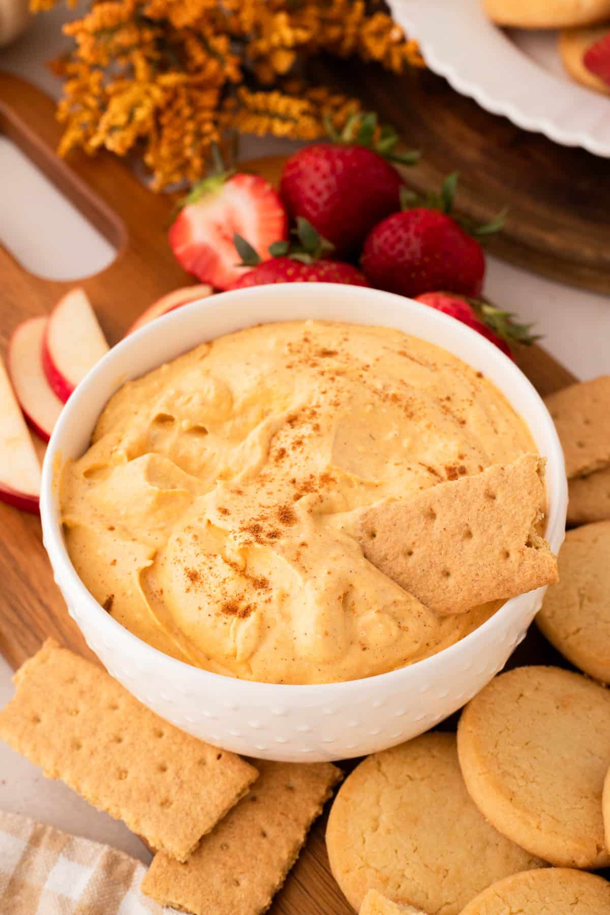 A bowl of pumpkin pie dip topped with a sprinkle of cinnamon, surrounded by graham crackers, cookies, apple slices, and strawberries on a wooden board.