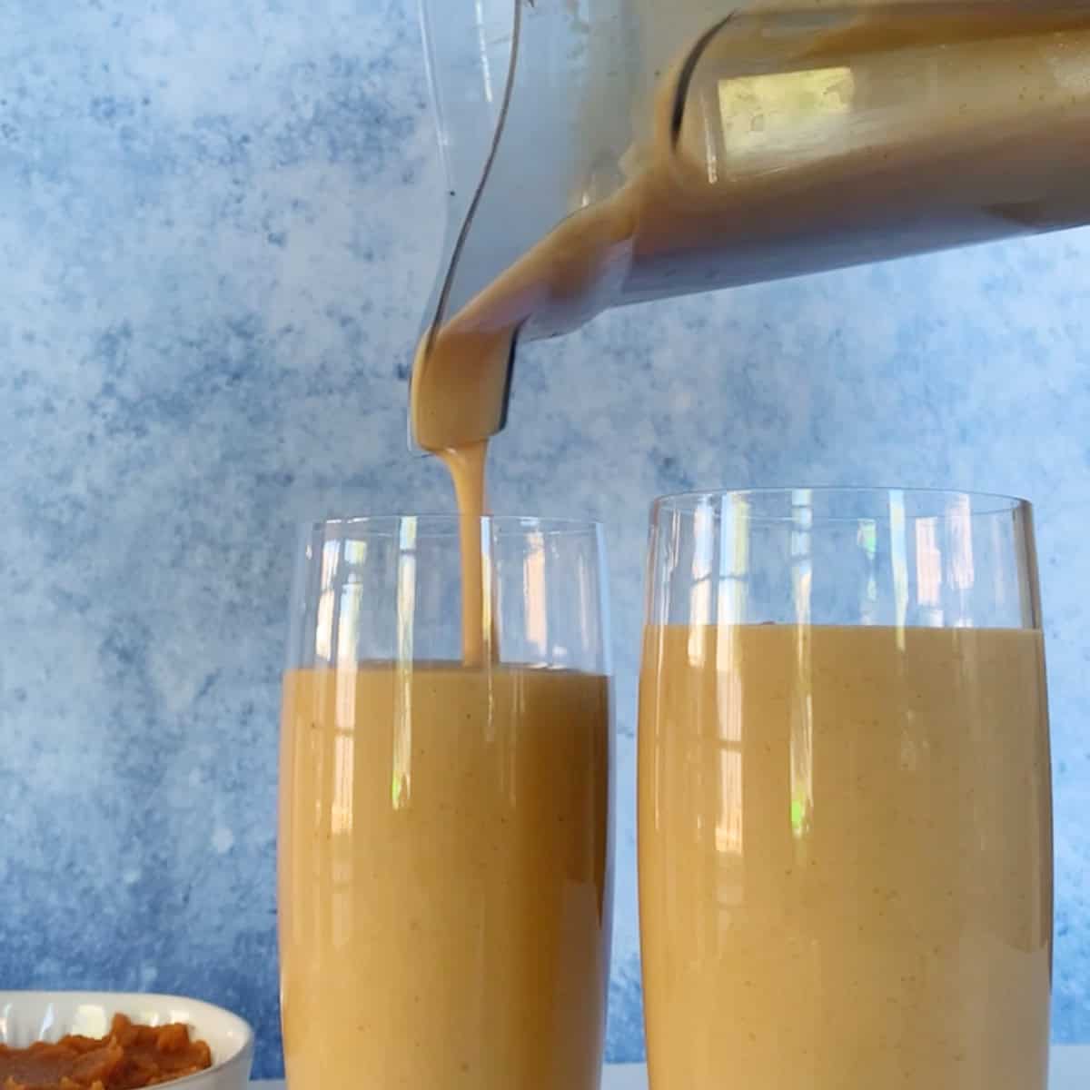 A blender is pouring a thick, light brown smoothie into a glass next to another filled glass, with a small bowl of food in the background.