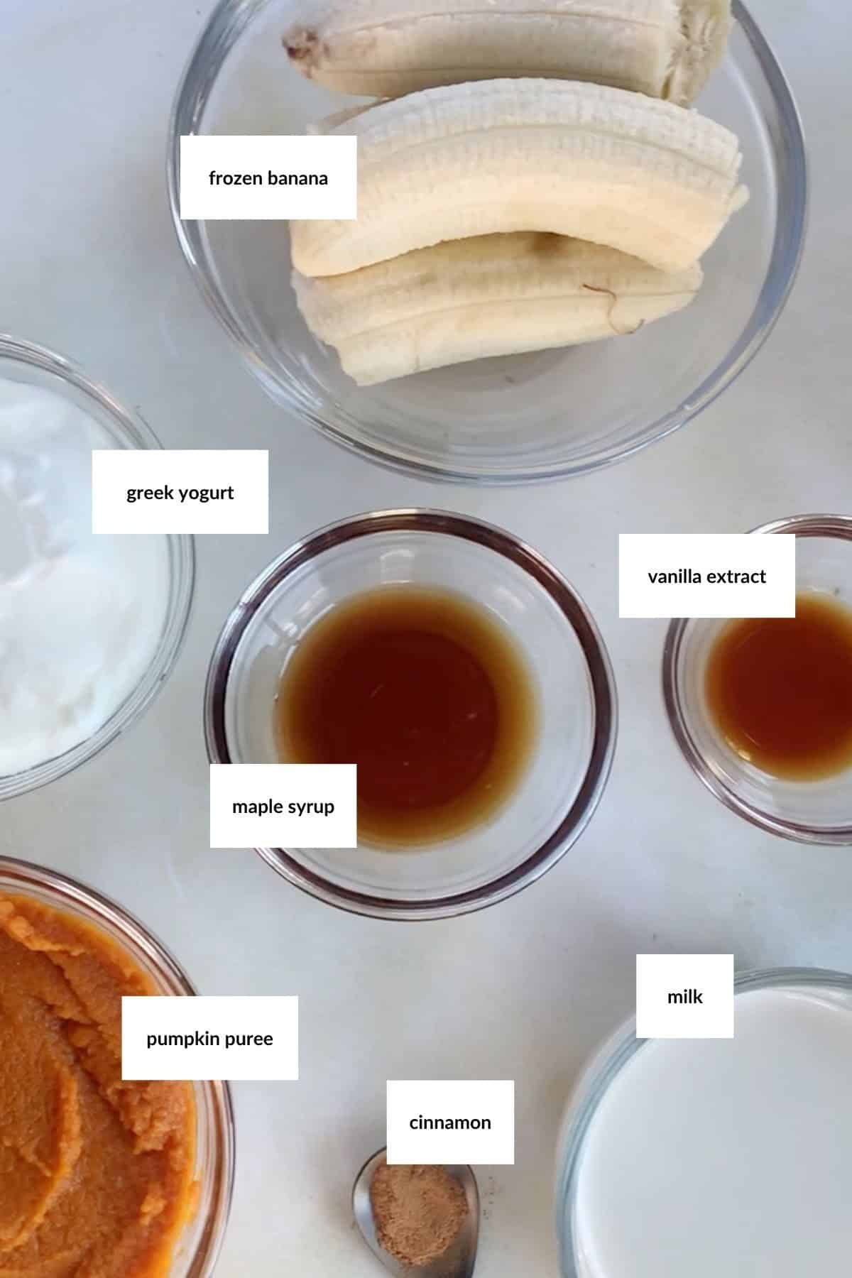 Bowls containing frozen banana, Greek yogurt, maple syrup, vanilla extract, pumpkin puree, cinnamon, and milk are arranged on a white surface, ready to be blended into a delicious pumpkin smoothie.