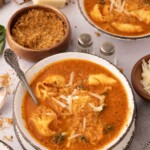 Bowl of tomato-based soup with tortellini and greens, topped with grated cheese, surrounded by bowls of breadcrumbs, cheese, and a wedge of parmesan.