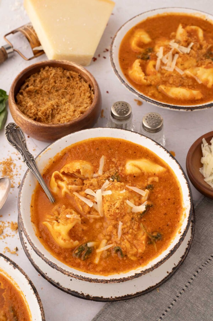 Bowl of tomato-based soup with tortellini and greens, topped with grated cheese, surrounded by bowls of breadcrumbs, cheese, and a wedge of parmesan.