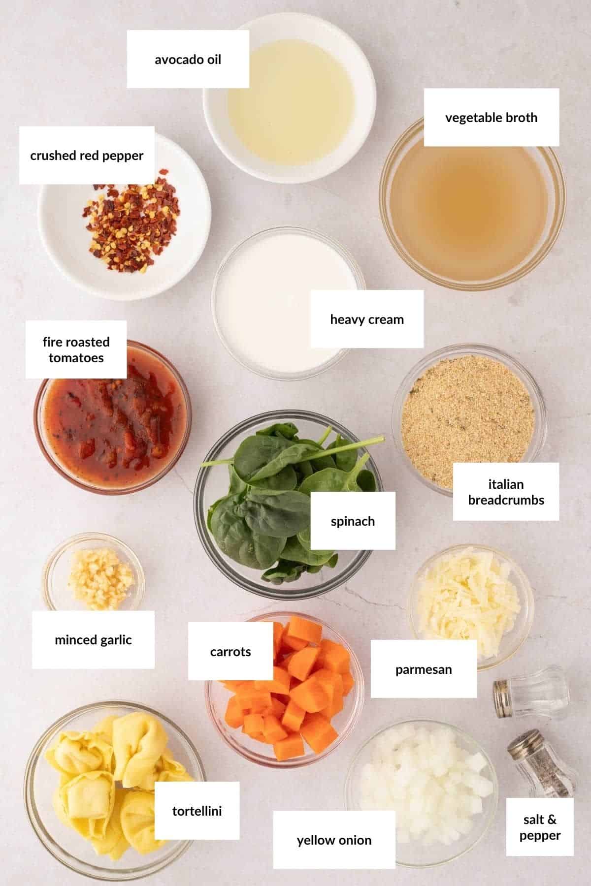 Various labeled ingredients for a spinach tortellini soup are arranged on a countertop, including avocado oil, vegetable broth, cream, breadcrumbs, tomatoes, and chopped vegetables.