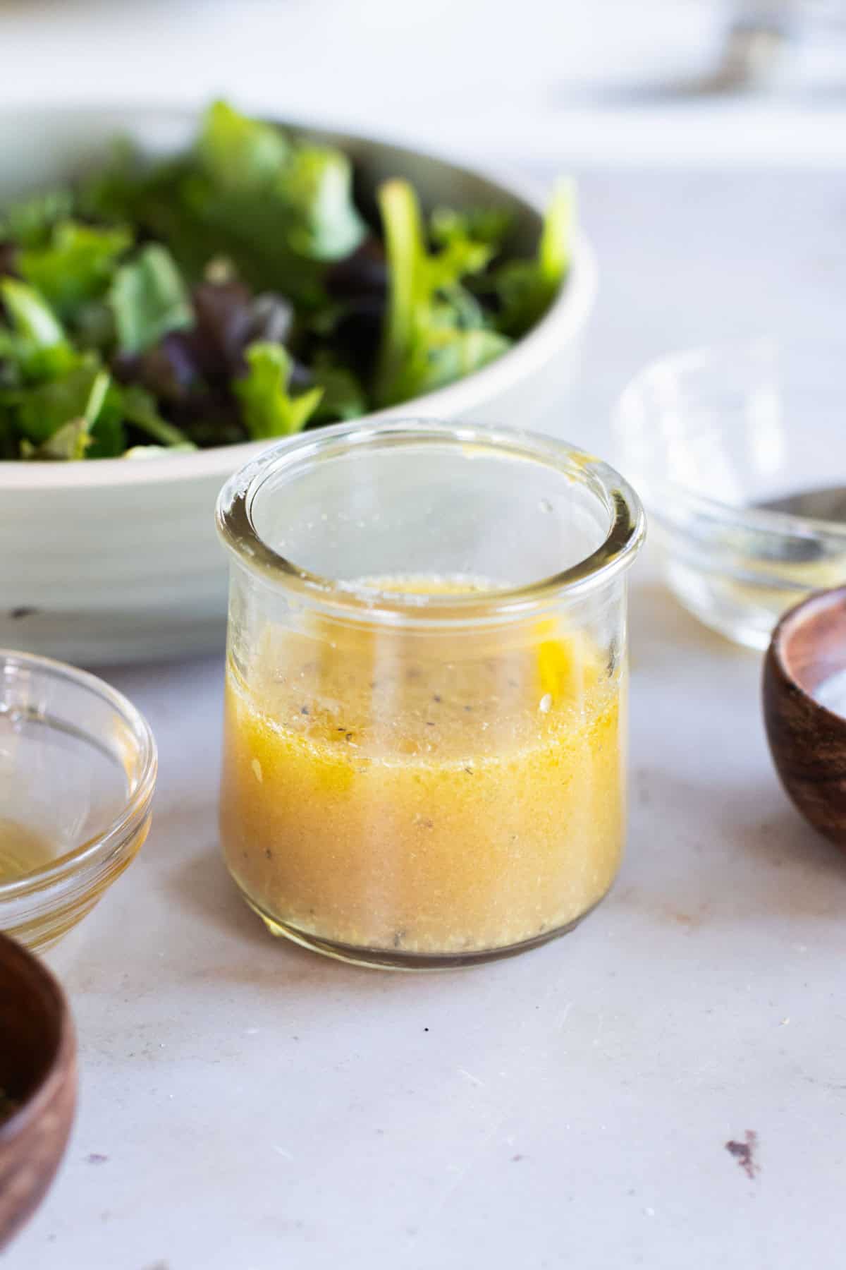 A small glass jar of vinaigrette dressing sits on a countertop near a bowl of mixed salad greens and several small bowls.