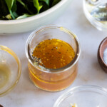 A small glass jar containing a golden liquid with black pepper and seasonings, surrounded by bowls of salt, garlic, and leafy greens.