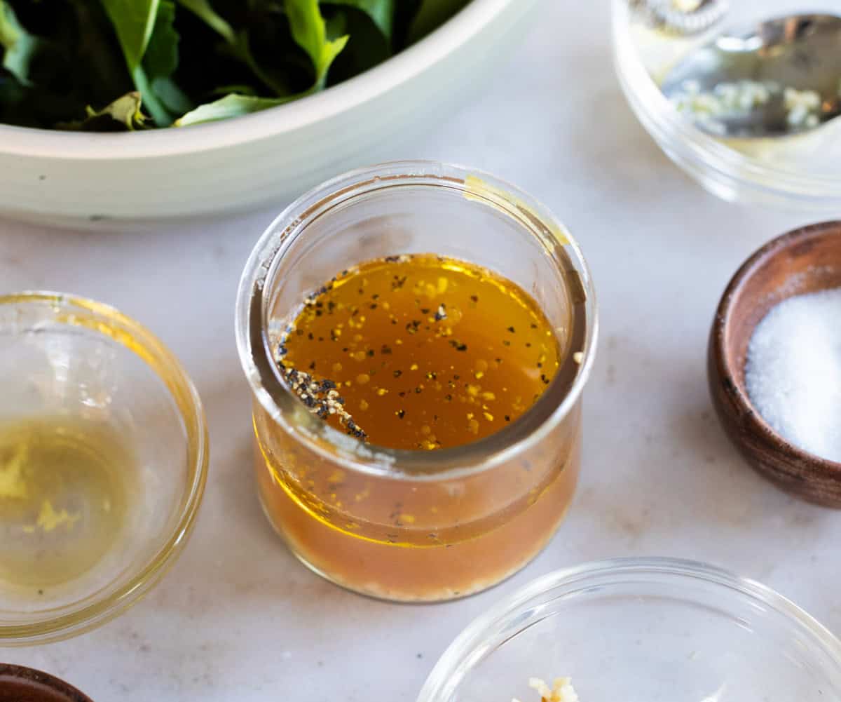 A small glass jar containing a golden liquid with black pepper and seasonings, surrounded by bowls of salt, garlic, and leafy greens.