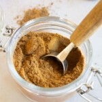 A glass jar filled with apple pie spice and a metal scoop with a wooden handle on a white surface.