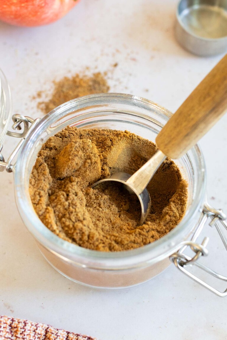 A glass jar filled with apple pie spice and a metal scoop with a wooden handle on a white surface.