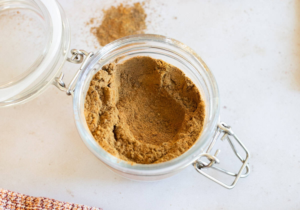 A glass jar with a metal clasp contains a light brown apple pie spice mixture, with some of the powder spilled on the white surface nearby.
