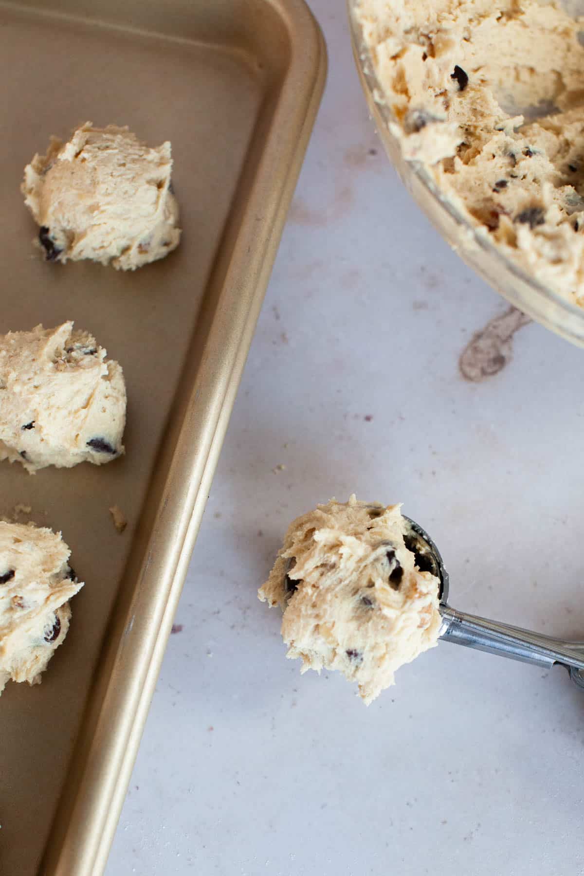 Scoops of chocolate chip cookie dough are placed on a baking sheet, with a mixing bowl and cookie scoop visible nearby.