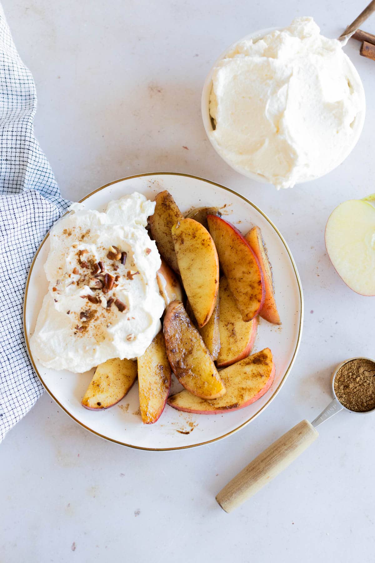 A plate with spiced apple slices and a large serving of whipped cream, garnished with nuts, sits beside a bowl of whipped cream, a measuring spoon with spice, and a cloth napkin.