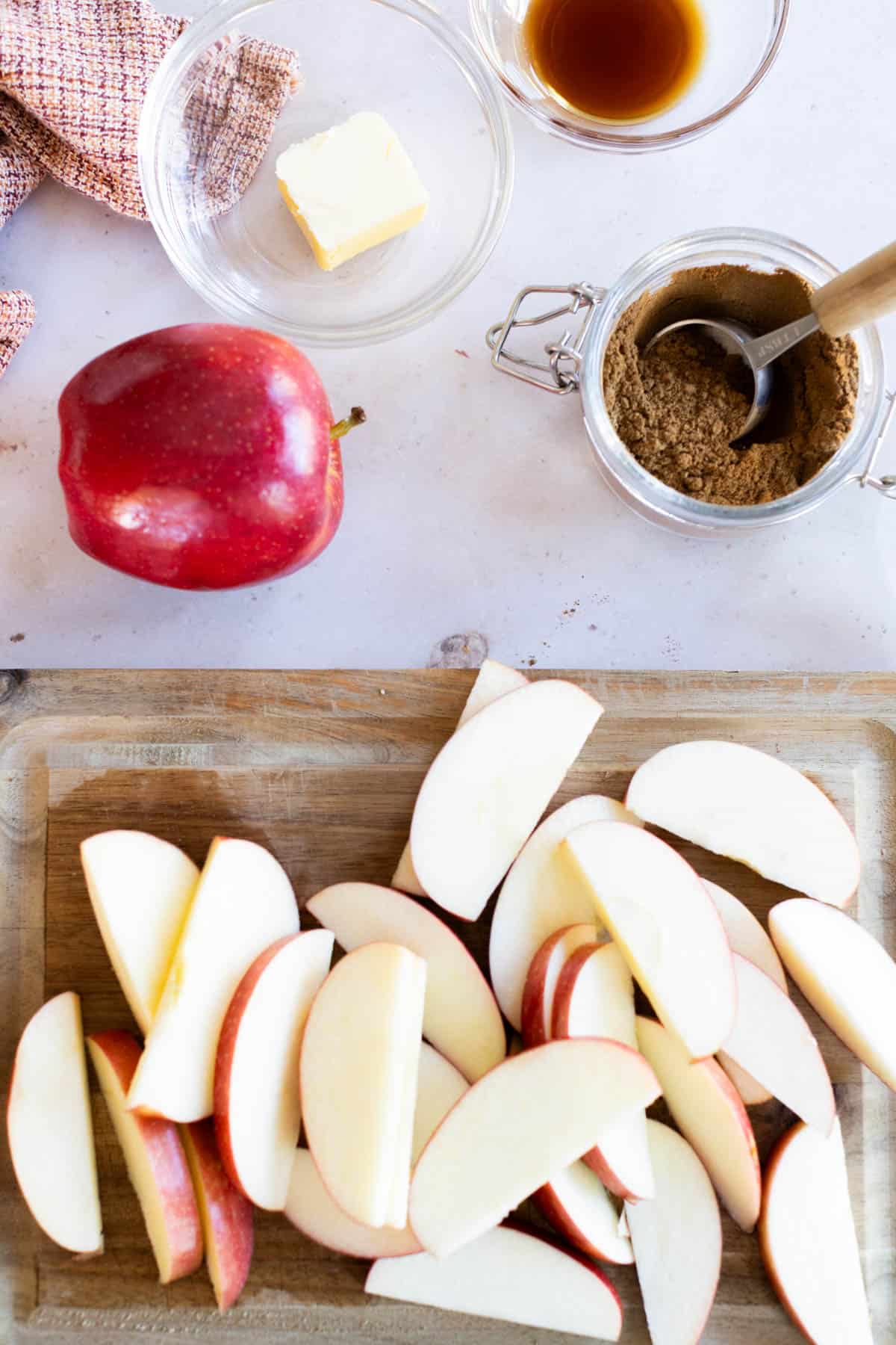 Sliced red apples on a cutting board, with a whole apple, a jar of cinnamon, a bowl of butter, and a bowl of vanilla extract on a countertop.