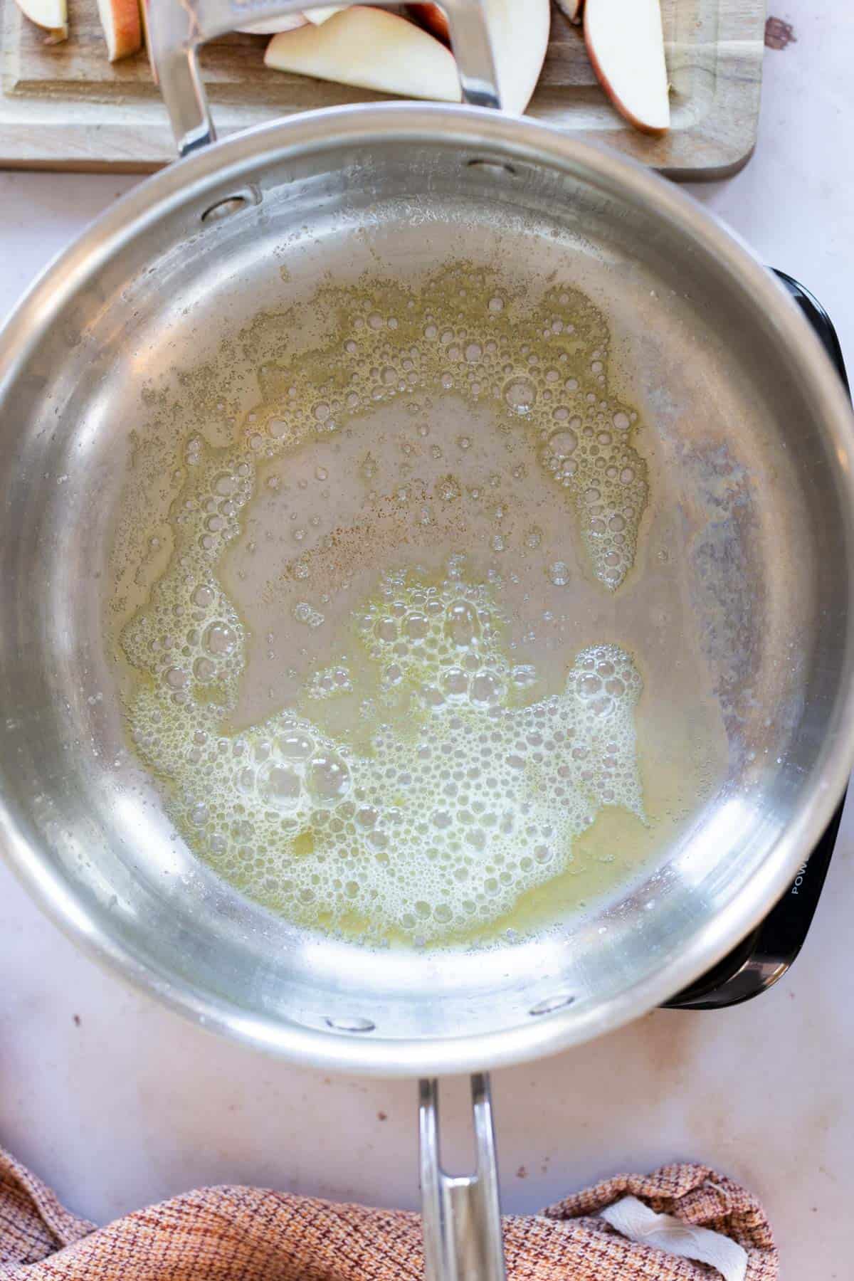 Melted butter with bubbles in a stainless steel pan, with sliced apples and a cutting board visible in the background.