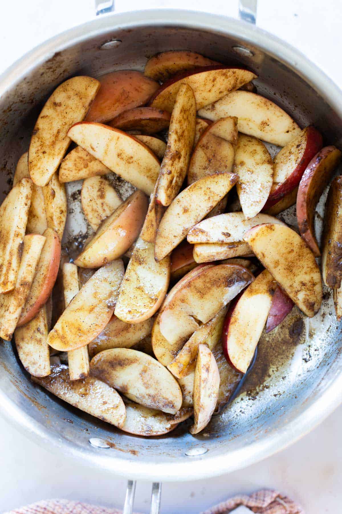 Sliced apples coated with cinnamon and seasoning are in a metal pot, ready for cooking or baking.