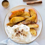 A plate of cinnamon-spiced apple slices with whipped cream, chopped nuts, a measuring cup of brown sugar, and cinnamon sticks on a white surface.