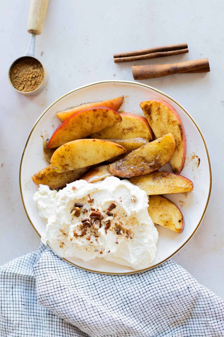 A plate of cinnamon-spiced apple slices with whipped cream, chopped nuts, a measuring cup of brown sugar, and cinnamon sticks on a white surface.