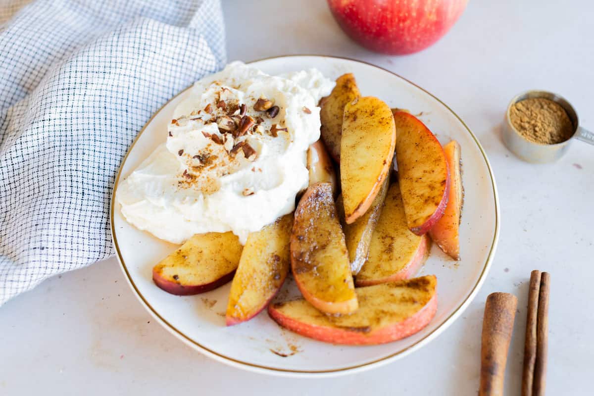 Plate with sliced baked apples topped with cinnamon, served with whipped cream and crushed nuts; a whole apple, cup of cinnamon, and cinnamon sticks nearby.