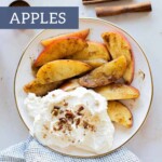 A plate of sliced cinnamon apples with whipped cream, garnished with chopped nuts, sits beside a blue towel and aromatic cinnamon sticks.