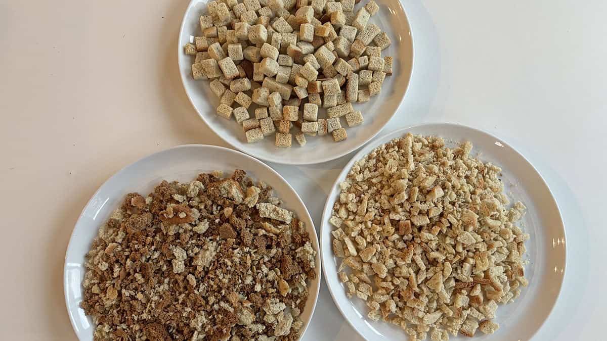 Three white plates, each containing different types of bread cubes and crumbs, are arranged on a white surface.