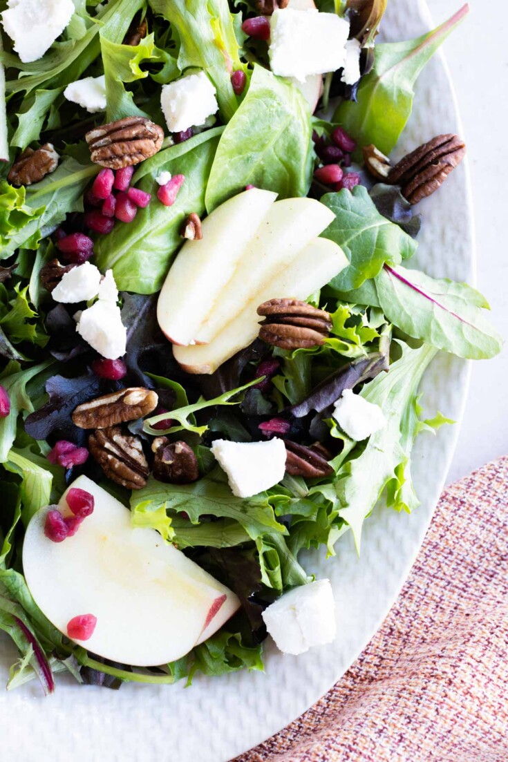 A salad with mixed greens, apple slices, pecans, pomegranate seeds, and crumbled white cheese on a white plate.