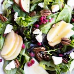 A salad with mixed greens, sliced apples, pecans, crumbled cheese, and pomegranate seeds on a white plate.