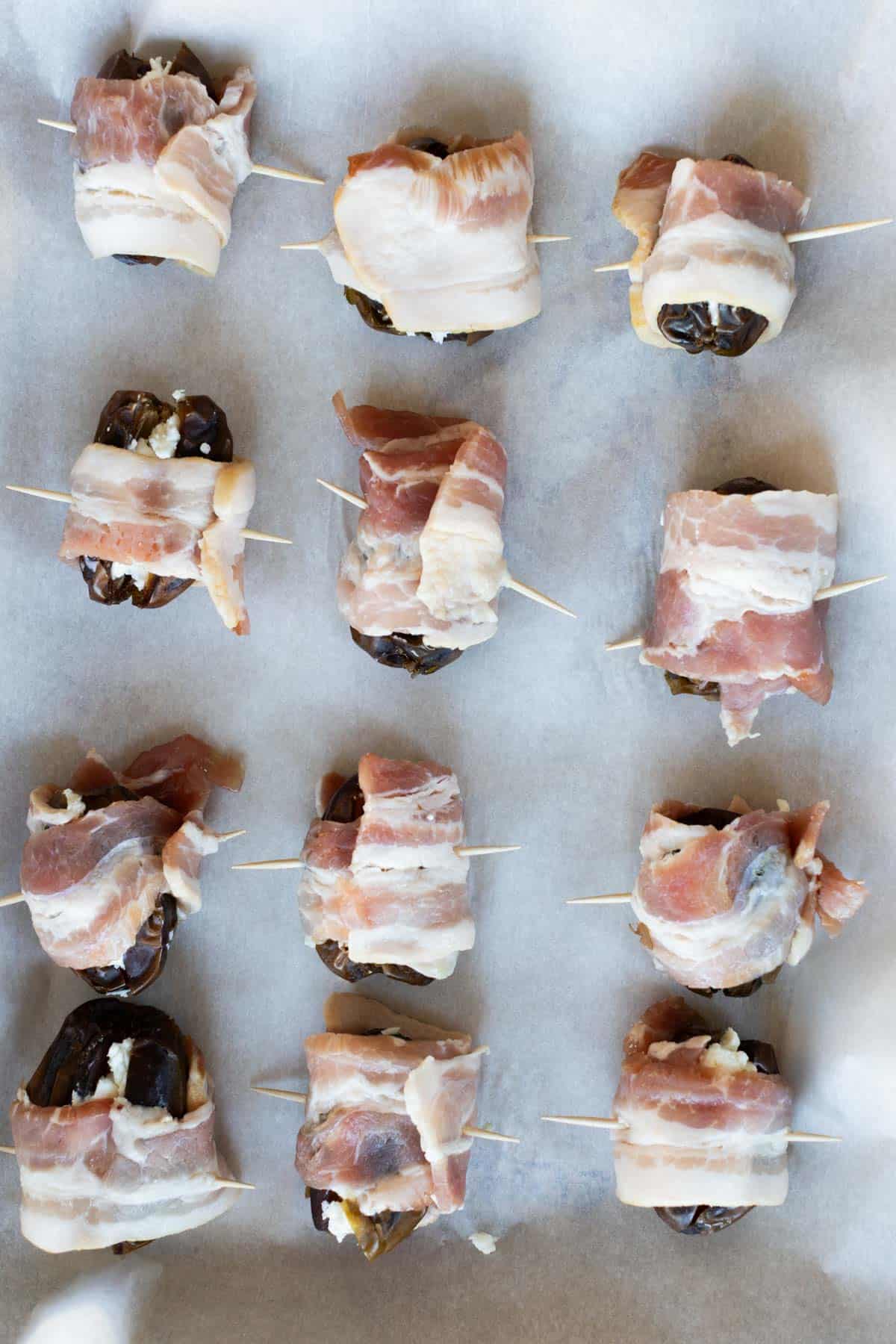 Twelve bacon-wrapped dates secured with toothpicks are arranged on a parchment-lined baking sheet, ready to be cooked.