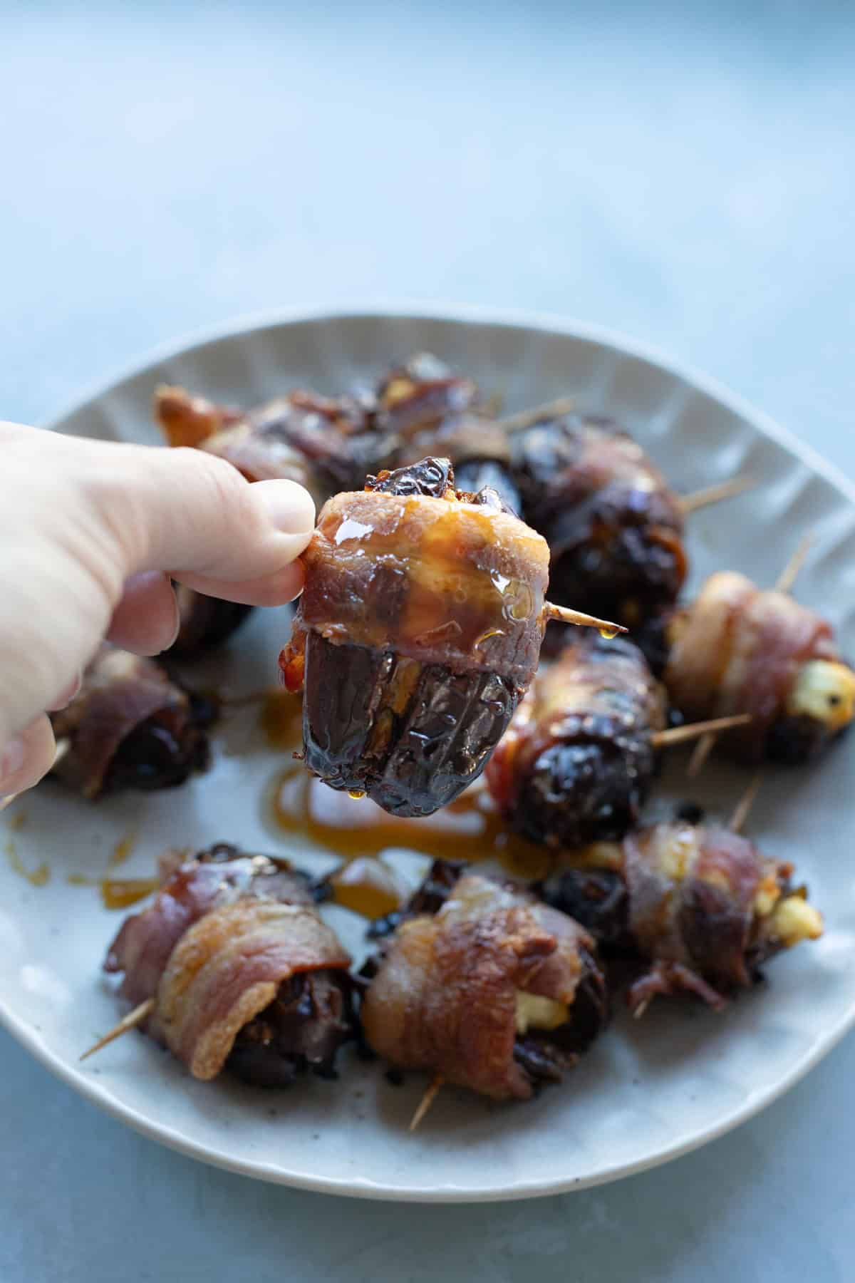 A hand holds a bacon-wrapped date with cheese, above a plate filled with more bacon-wrapped dates secured with toothpicks.