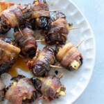 A plate of bacon-wrapped dates secured with toothpicks, some slightly caramelized and stuffed, arranged on a white dish.