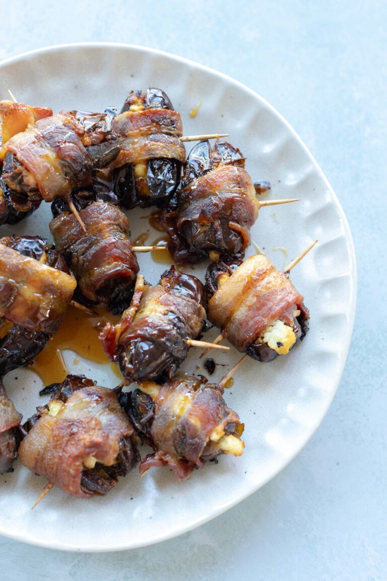 A plate of bacon-wrapped dates secured with toothpicks, some slightly caramelized and stuffed, arranged on a white dish.