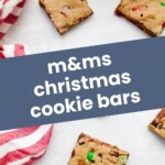 Top-down view of four M&M Christmas Cookie Bars on a white surface next to a red and white striped cloth. Text overlay reads "m&ms christmas cookie bars" and "joyfullymad.com".
