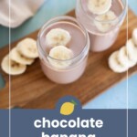 Two glasses of chocolate banana smoothie topped with banana slices, placed on a wooden board with additional banana slices beside them.