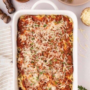 A baked cream cheese spaghetti casserole topped with melted cheese and herbs in a white baking dish, surrounded by plates, forks, seasonings, and a kitchen towel.