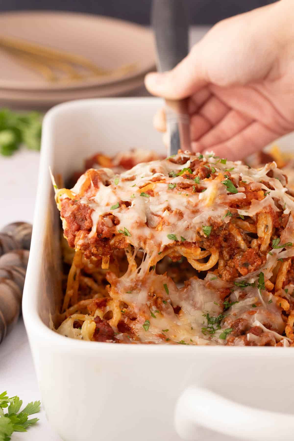 A hand serves a portion of cream cheese baked spaghetti with meat sauce from a white casserole dish, garnished with chopped herbs.
