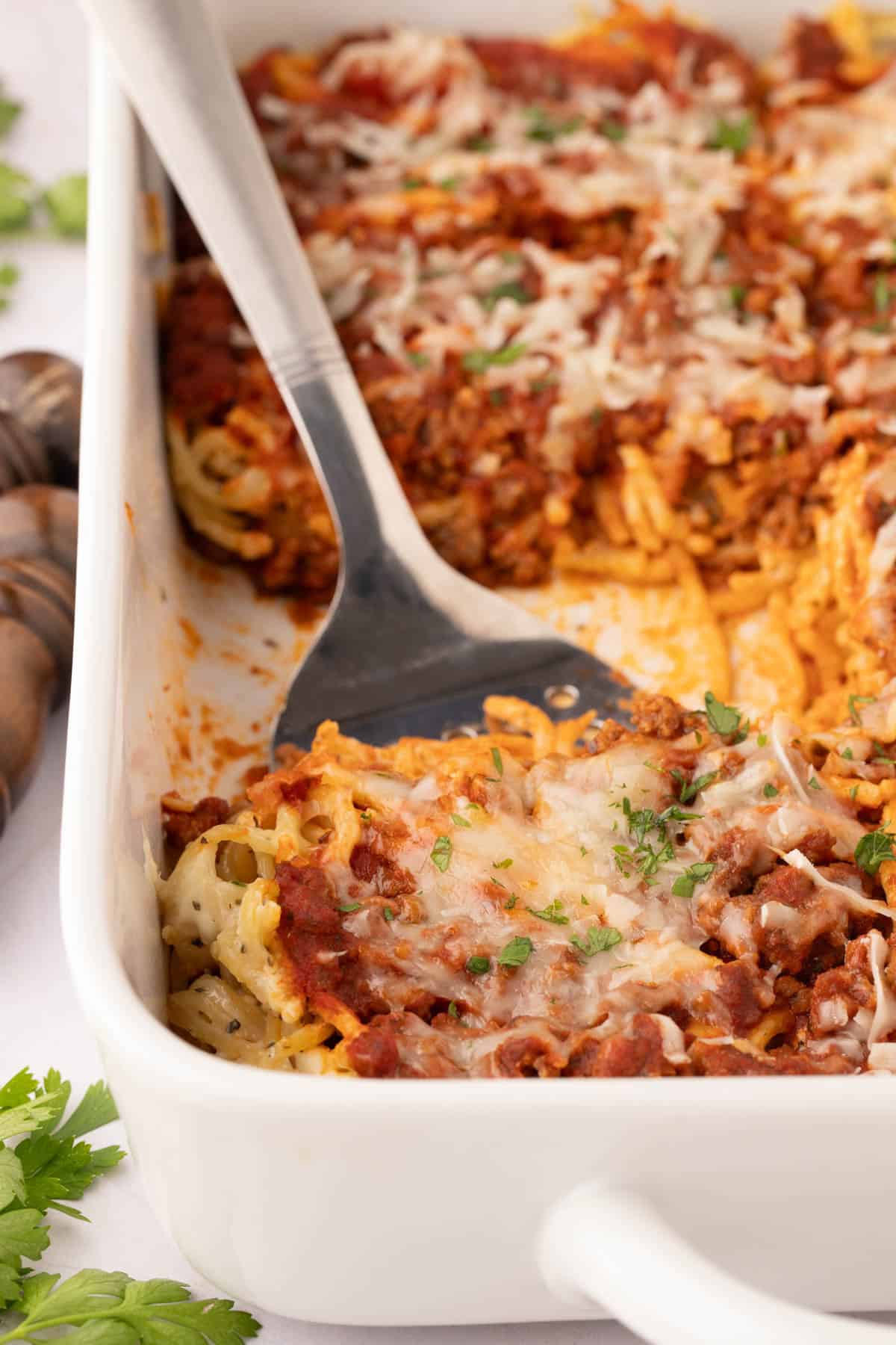 A dish filled with cream cheese casserole spaghetti topped with melted cheese and tomato sauce, with a serving spatula lifting a portion. Fresh herbs are sprinkled on top.