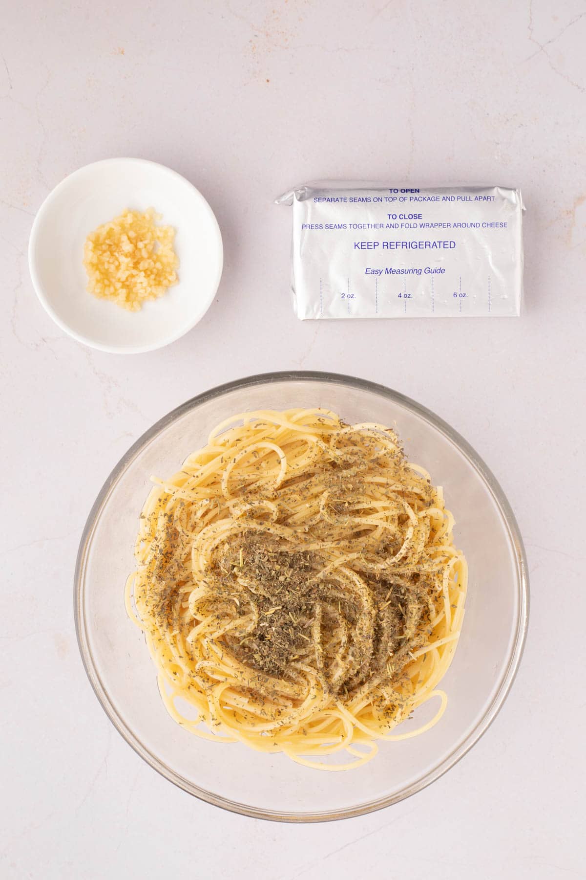 A bowl of cooked spaghetti pasta seasoned with herbs, a small dish of minced garlic, and a wrapped block of cream cheese on a light countertop.