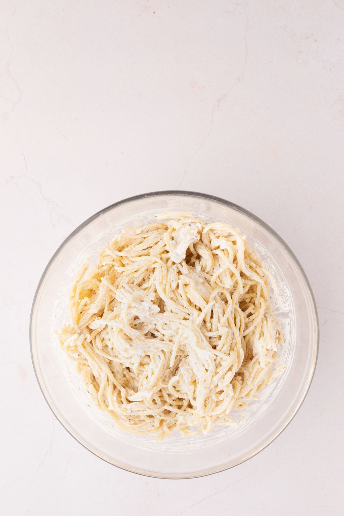 A clear glass bowl filled with cooked spaghetti pasta mixed with a cream cheese, placed on a light-colored surface.