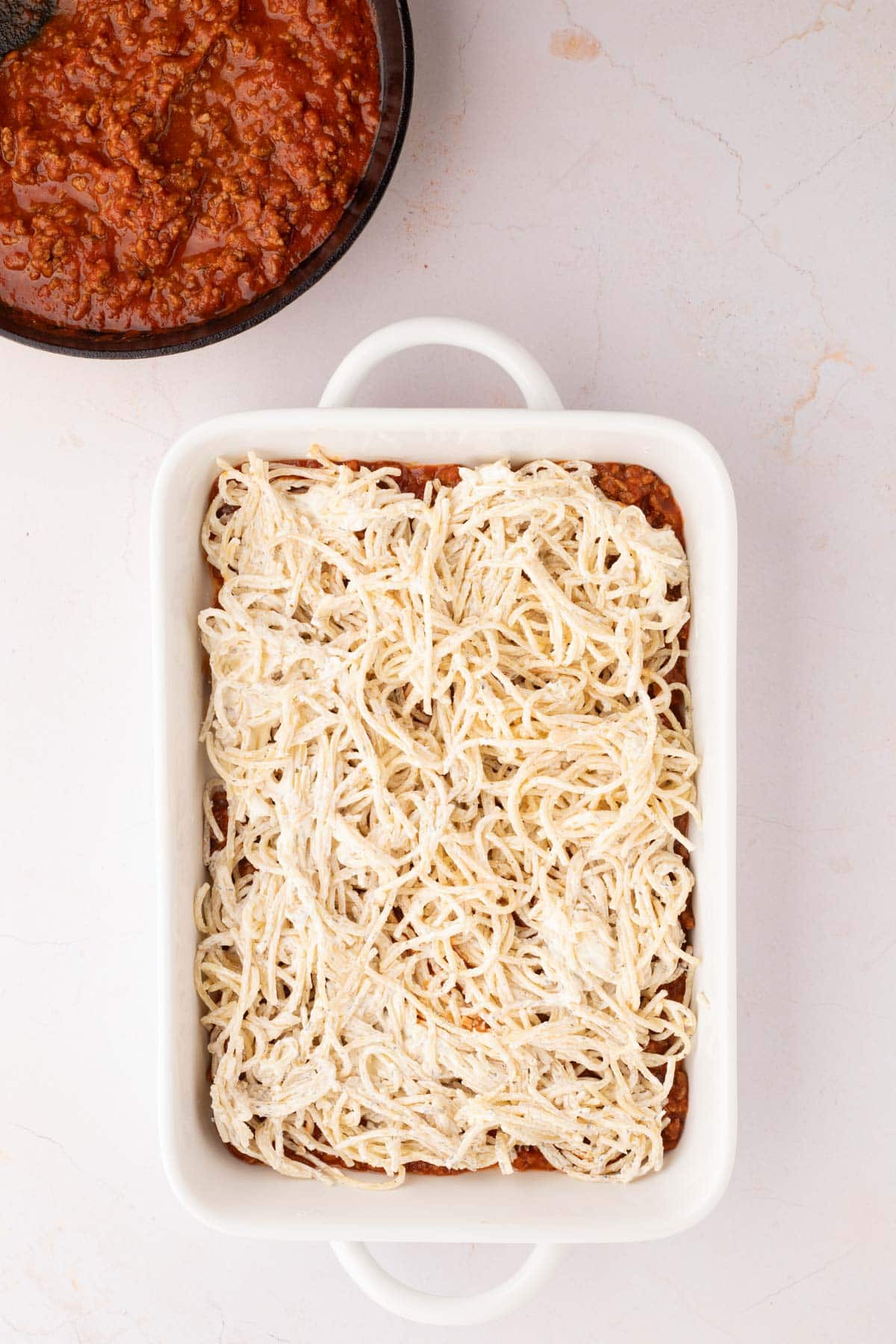 A white baking dish filled with spaghetti mixed with a cream cheese sauce, placed next to a bowl of red meat sauce on a light surface.