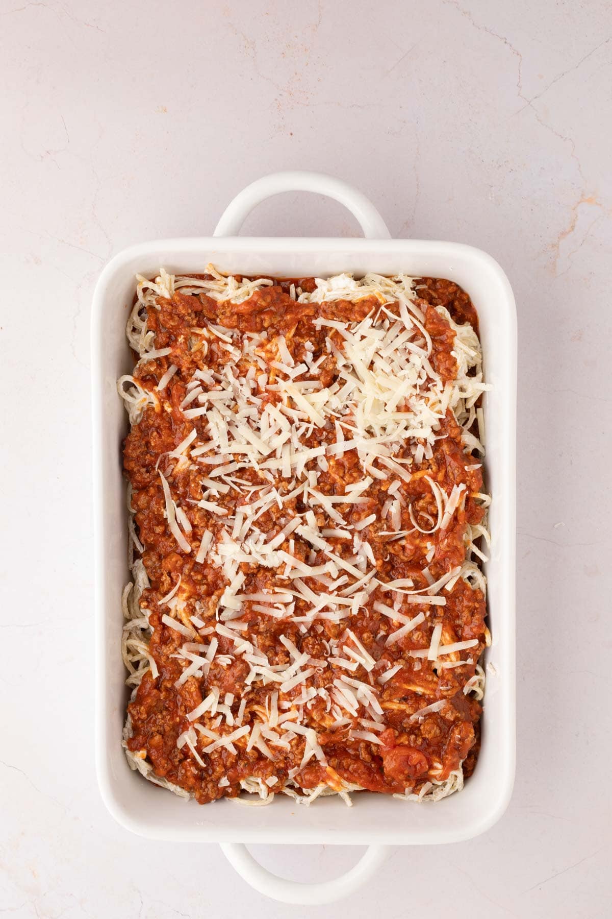 A rectangular white baking dish filled with shredded cheese and meat sauce over noodles, prepared for baking.