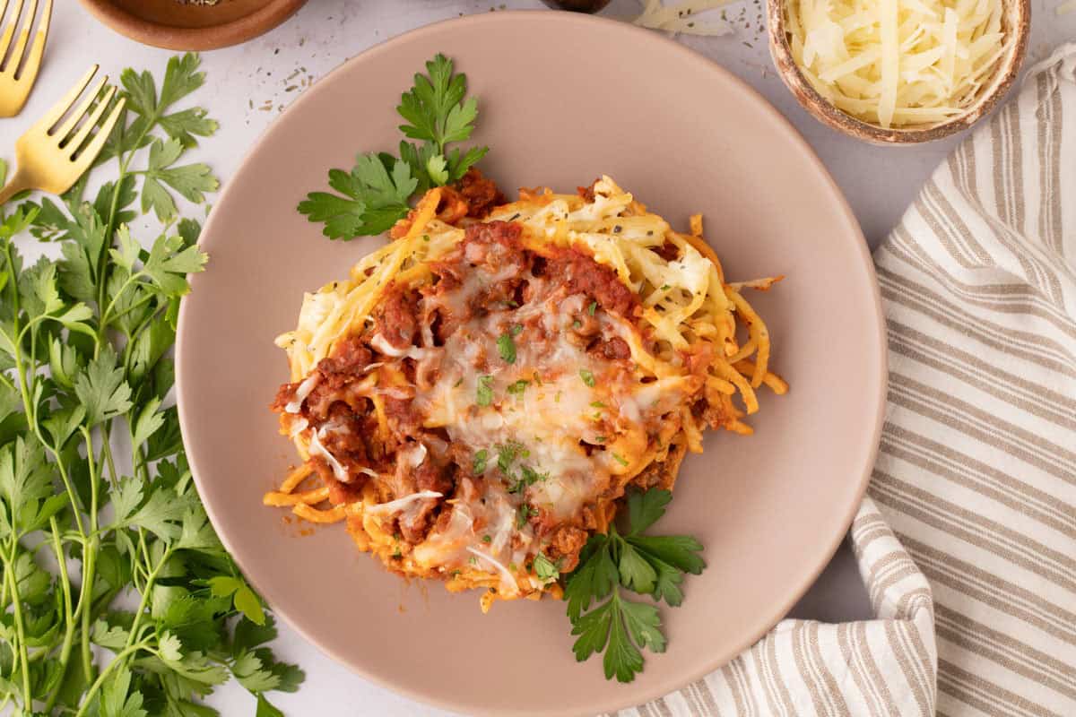 A plate of cream cheese spaghetti casserole with tomato sauce and melted cheese, garnished with parsley, is placed on a table next to shredded cheese, parsley, and a striped napkin.