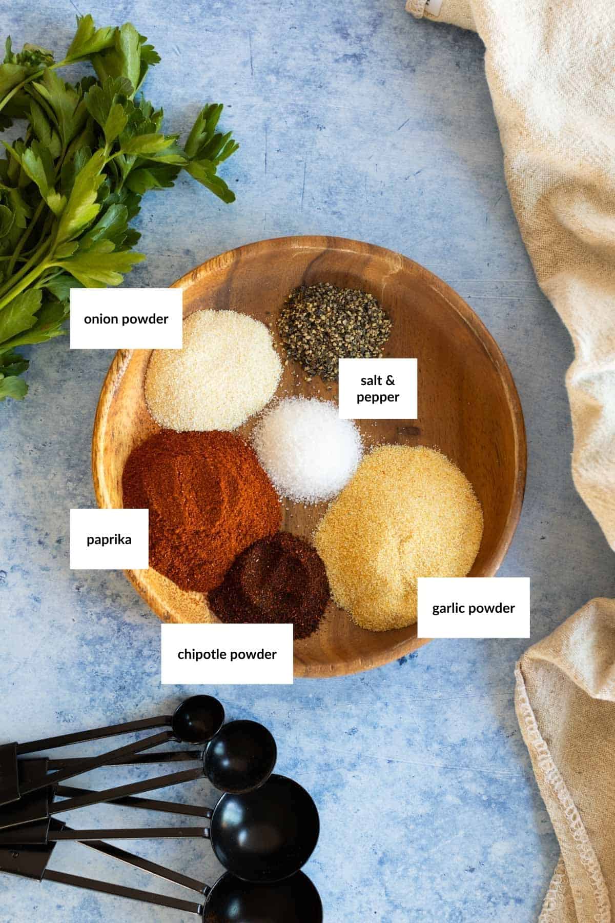 A wooden bowl containing paprika, chipotle powder, onion powder, garlic powder, salt, pepper, surrounded by parsley, measuring spoons, and a beige cloth on a blue surface. Labels identify each spice.