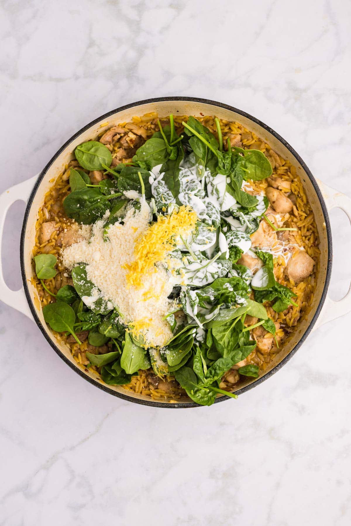 A white pot filled with rice, spinach, pieces of chicken, grated cheese, lemon zest, and creamy sauce on a marble surface.
