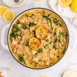 A pot of creamy orzo with spinach, chicken, and two grilled lemon halves, surrounded by plates, utensils, lemon slices, and seasonings on a white surface.