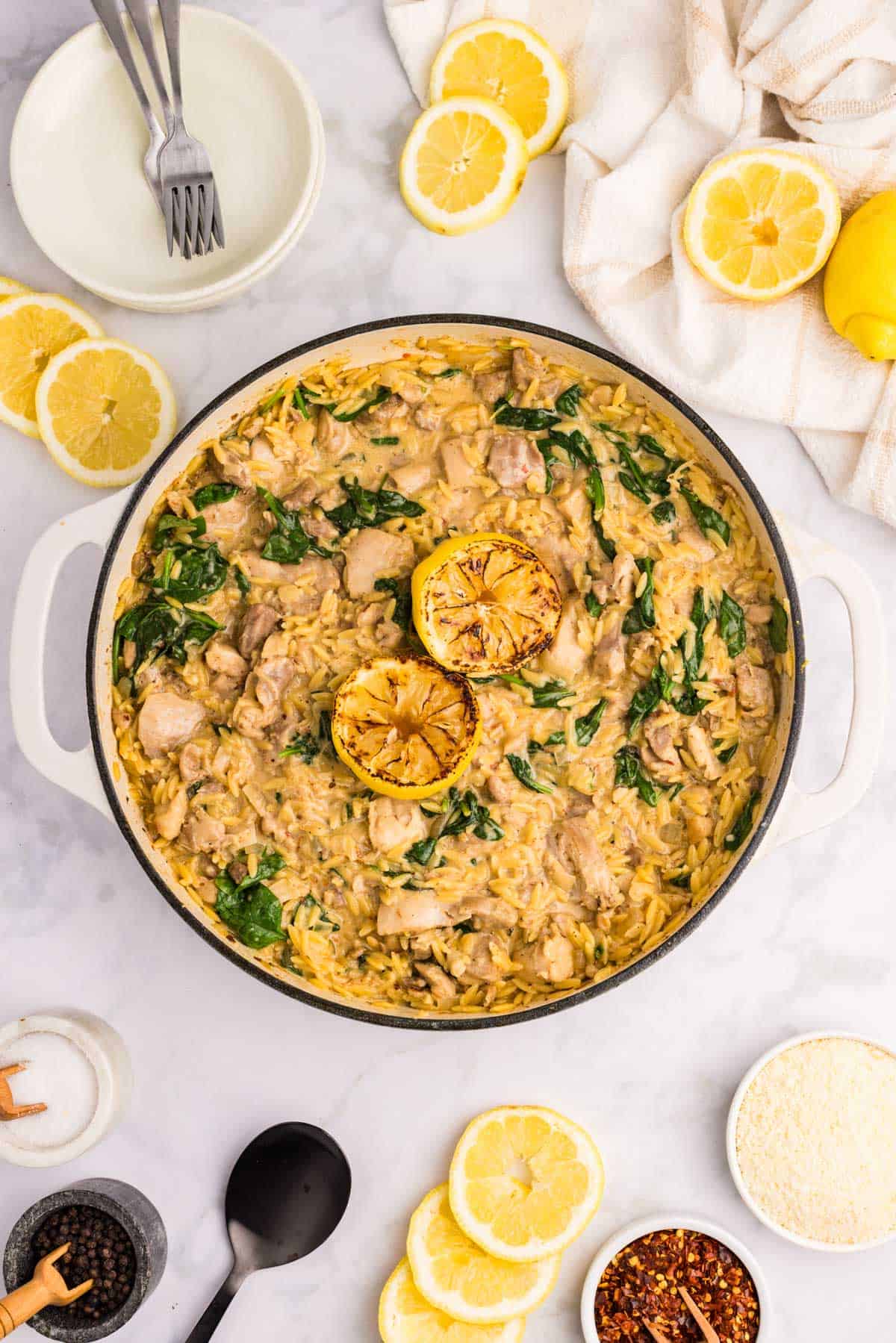 A pot of creamy orzo with spinach, chicken, and two grilled lemon halves, surrounded by plates, utensils, lemon slices, and seasonings on a white surface.