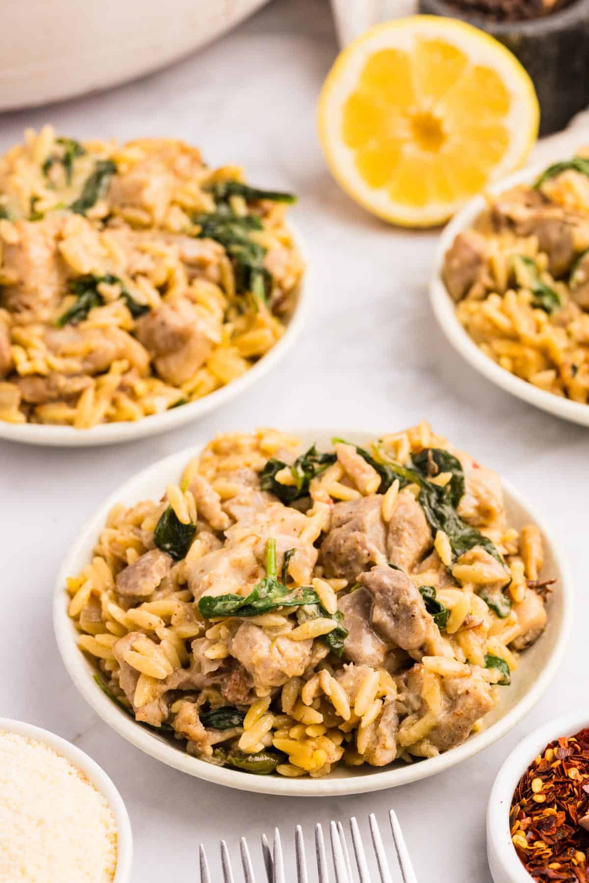 Bowls of creamy orzo pasta with pieces of chicken and spinach, surrounded by a lemon half, grated cheese, and red pepper flakes on a white surface.